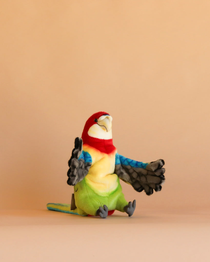 The Parrot Puppet, with its red head, yellow belly, green tail, and blue wings, sits upright against a plain beige background. Carefully hand sewn, this colorful puppet stands out among plush animals for its soft texture and realistic features.