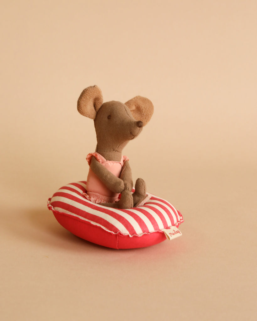 A small brown fabric mouse, crafted from cotton fabric, sits on a Maileg Beach Raft, Small Mouse - Red stripe against a beige background. The mouse is wearing a pink outfit and is positioned in a relaxed, seated pose. The overall scene has a warm, minimalist aesthetic reminiscent of baby mice sizes.