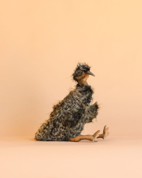 A fluffy, brown and black Emu Puppet with orange legs sits on a beige background, facing left with its beak slightly open—capturing the lifelike charm of HANSA animals and their hand-sewn stuffed animals.