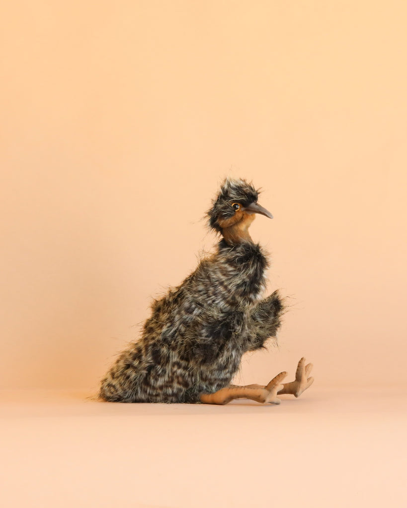 A fluffy, brown and black Emu Puppet with orange legs sits on a beige background, facing left with its beak slightly open—capturing the lifelike charm of HANSA animals and their hand-sewn stuffed animals.