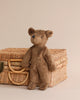 A Senger Naturwelt Bear Stuffed Animal sitting on a wicker picnic basket with a beige background. The bear, created with sustainable practices, has a slight tilt to its head and blue button eyes.