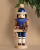 The Christian Ulbricht Collectible Wooden Nutcracker - Gingerbread Vendor, a limited-edition piece crafted in Germany, depicts a soldier adorned in a blue winter hat and scarf. He holds a tray of gingerbread cookies while standing under a green pine branch set against a light brown background, encapsulating the charm of traditional craftsmanship.