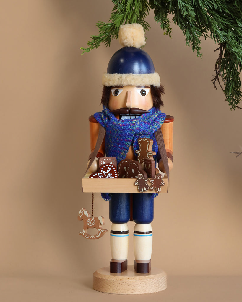The Christian Ulbricht Collectible Wooden Nutcracker - Gingerbread Vendor, a limited-edition piece crafted in Germany, depicts a soldier adorned in a blue winter hat and scarf. He holds a tray of gingerbread cookies while standing under a green pine branch set against a light brown background, encapsulating the charm of traditional craftsmanship.