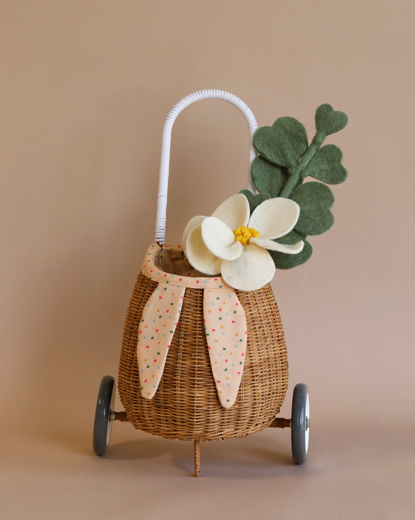 A decorative Rattan Bunny Luggy With Lining – Gumdrop on wheels with a white handle, adorned with a polka dot fabric bow and a faux white flower with green leaves, set against a plain beige background.