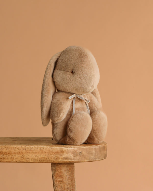 The Maileg Plush Bunny, Medium - Cream Peach, with floppy ears and a small ribbon around its neck, sits on a wooden stool against a plain beige background.