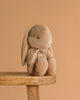 The Maileg Plush Bunny, Medium - Cream Peach, with floppy ears and a small ribbon around its neck, sits on a wooden stool against a plain beige background.