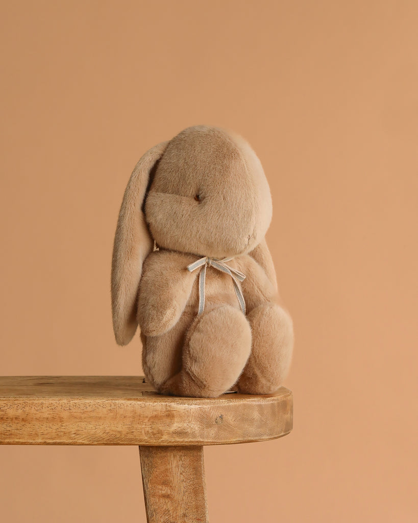 The Maileg Plush Bunny, Medium - Cream Peach, with floppy ears and a small ribbon around its neck, sits on a wooden stool against a plain beige background.