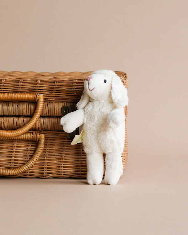 A Senger Naturwelt Stuffed Animal - Baby Sheep, handmade in Germany and standing in front of a wicker basket on a soft beige background.