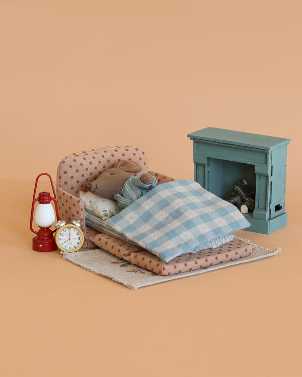 The Maileg Big Brother Family Room Set features a miniature bedroom scene with a small bed, blue checkered blanket, Maileg Big Brother Mouse figure, teal fireplace, red lantern, and white alarm clock—ideal for dollhouse accessories.