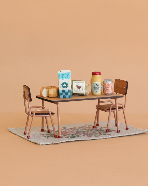 A Maileg Dining Table Set sits on a Maileg Flower Rug, with miniature bread, milk, eggs, honey, and jam arranged on the table against a plain peach background.