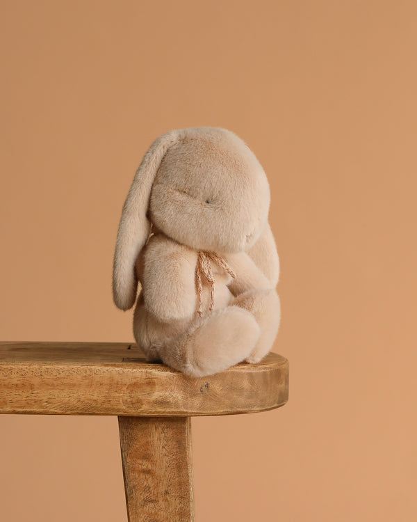The Maileg Plush Bunny, Small - Latte, with gently drooping long ears and closed eyes, sits quietly on a wooden stool against a warm beige background, its soft paws resting in its lap.