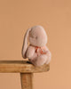 The Maileg Plush Bunny, Small - Powder, with a light pink hue and a bow around its neck, rests on the edge of a wooden stool against a plain peach background, its soft fabric inviting gentle hugs.