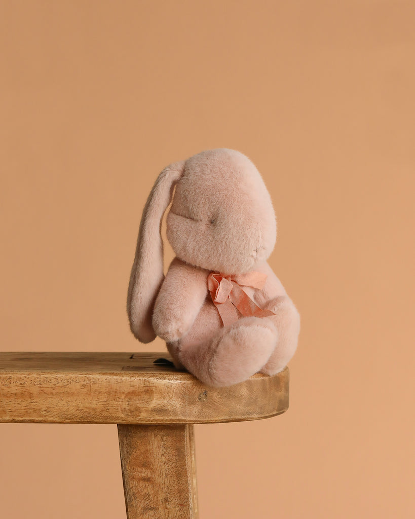 The Maileg Plush Bunny, Small - Powder, with a light pink hue and a bow around its neck, rests on the edge of a wooden stool against a plain peach background, its soft fabric inviting gentle hugs.
