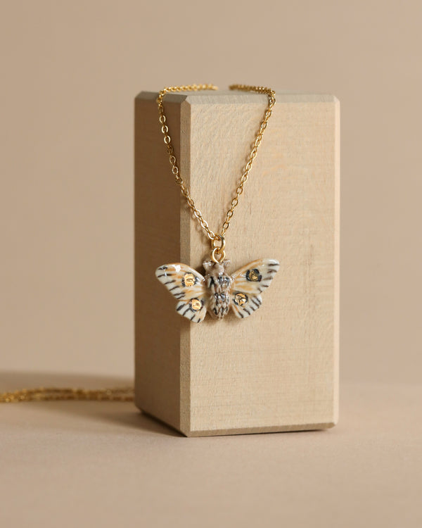  A Solar Moth Necklace with detailed wings hangs from a beige jewelry box against a soft beige background, emphasizing its elegant design.