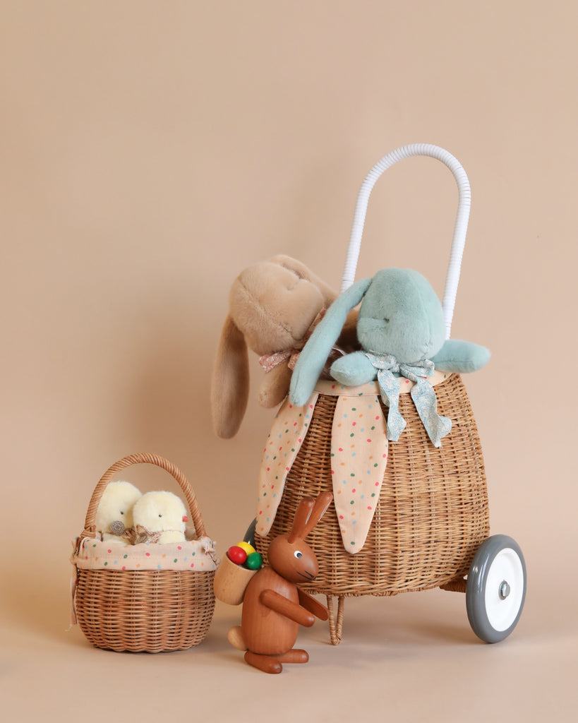 A charming display of a large Rattan Berry Basket With Lining – Gumdrop on wheels adorned with two plush rabbits, next to a small storage basket filled with colorful eggs, all against a soft beige background.