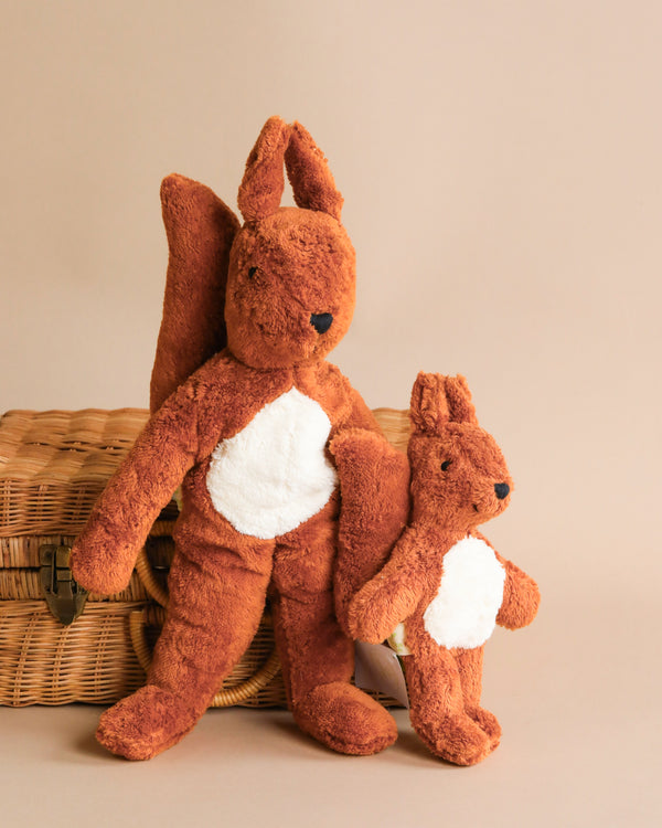 Two plush squirrel toys, one large and one small, crafted from organically grown cotton, leaning against a wicker basket on a light brown background.
