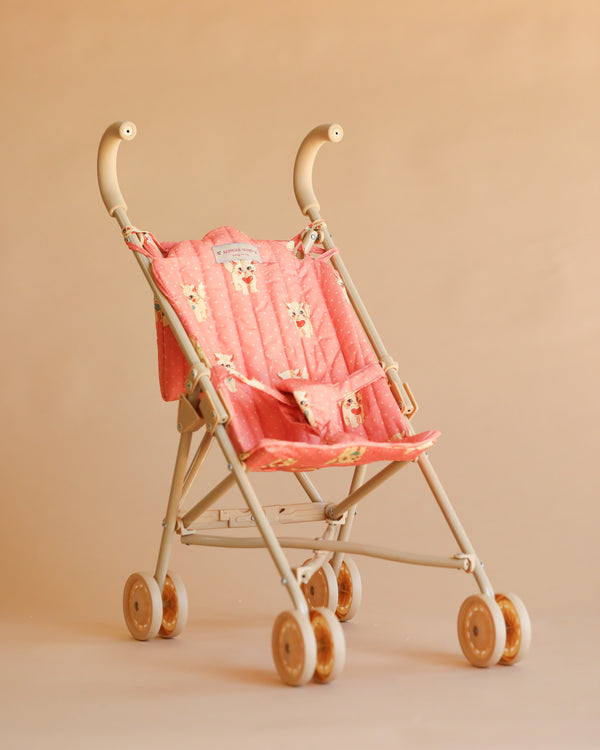 A Konges Sloejd Doll Stroller in the Kitty Ciao Miao Rose design, featuring a beige and pink color scheme with cat prints on the seat and double wheels, set against a plain light brown background.