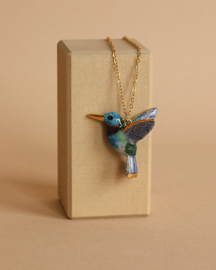 A delicate Hummingbird Necklace with a pendant shaped like a small, colorful hummingbird is displayed against a plain beige background. The bird, featuring a blue head, green body, and vibrant hand-painted feather wings, is suspended on a thin 24k gold plated chain draped over a wooden rectangular stand.