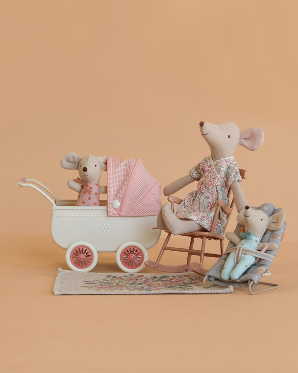 The Maileg Nursery Set features three fabric toy mice arranged as a family with nursery furniture—a rocking chair, bouncer, and baby carriage with pink blanket—displayed on a patterned rug against a peach background.