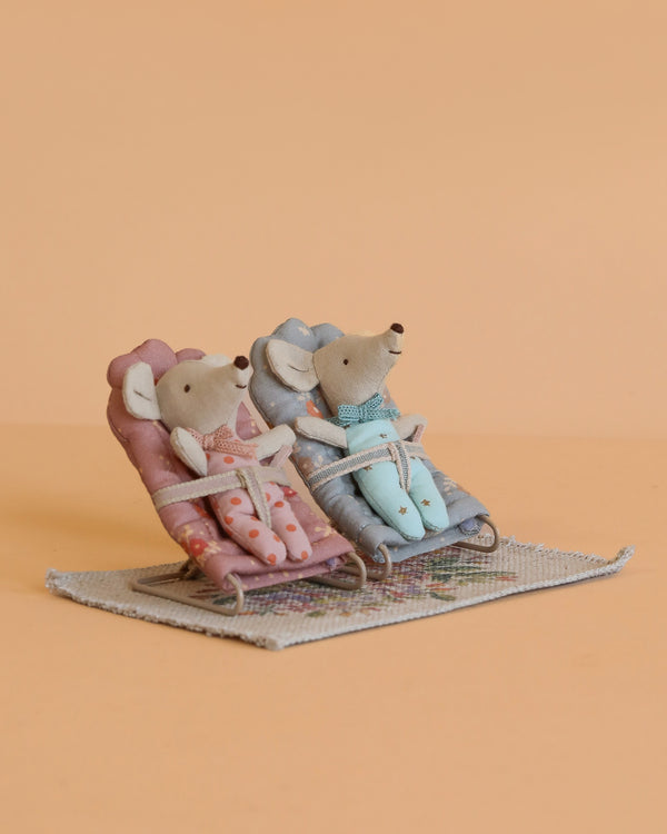 The Maileg Baby Twins with Babysitters Set features two baby mice in bathrobes relaxing on pink and blue chairs atop a dollhouse rug, all set against a plain peach background.