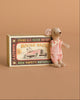 A Maileg Family Celebration Set features a charming small mouse doll dressed in pink attire, nestled beside a vintage-style matchbox labeled “Mouse Race,” adorned with a race car illustration on a brown background—an adorable addition to any Maileg mice in matchbox collection.