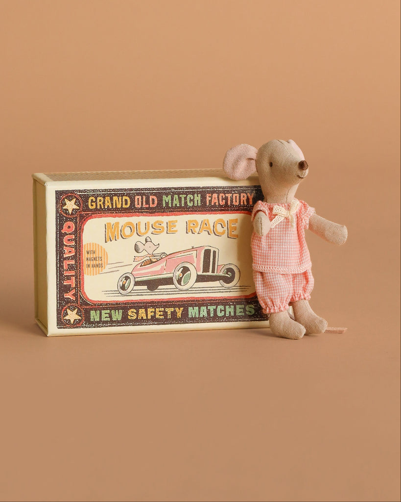 A Maileg Family Celebration Set features a charming small mouse doll dressed in pink attire, nestled beside a vintage-style matchbox labeled “Mouse Race,” adorned with a race car illustration on a brown background—an adorable addition to any Maileg mice in matchbox collection.