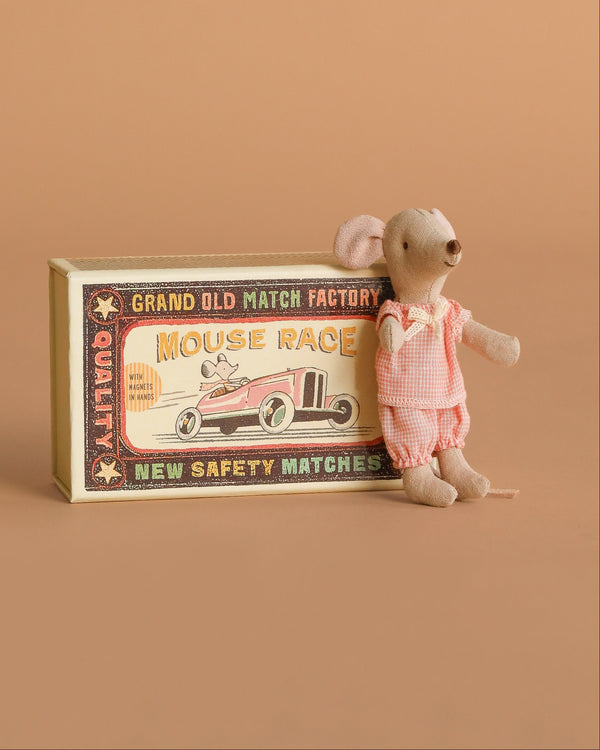 The Maileg Little Sister Mouse in pink clothes stands beside her vintage-style “Mouse Race” matchbox, which shows a mouse driving a race car, all set against a solid light brown background.