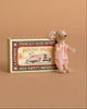 The Maileg Little Sister Mouse in pink clothes stands beside her vintage-style “Mouse Race” matchbox, which shows a mouse driving a race car, all set against a solid light brown background.