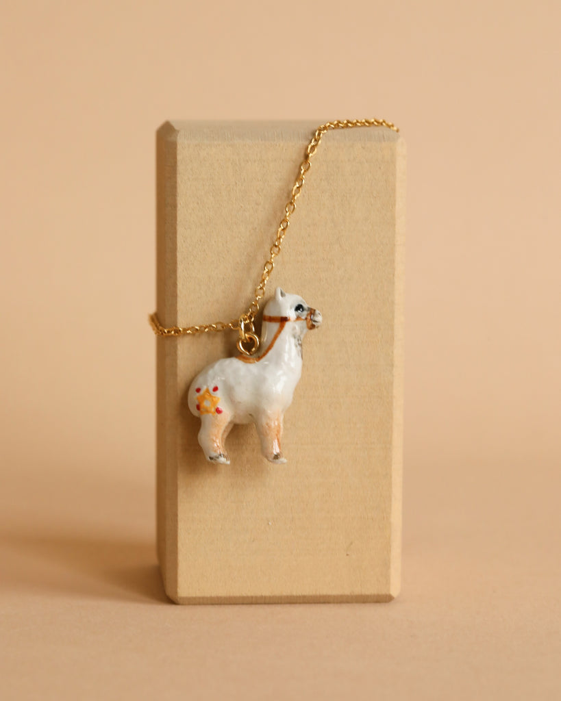 A handcrafted jewelry piece, this alpaca necklace boasts a white pendant adorned with colorful floral designs. It is elegantly draped over a plain, rectangular beige box. The background is a soft, matching beige, enhancing the 24k gold chain's detailing.