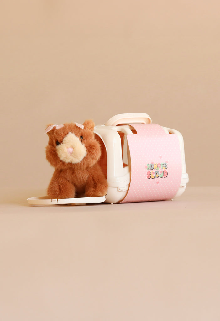A plush toy cat from Konges Sloejd’s “Take Care of Me Animals” series sits halfway inside a small white plastic pet carrier with a pink polka-dot door, inviting you to take care of it.