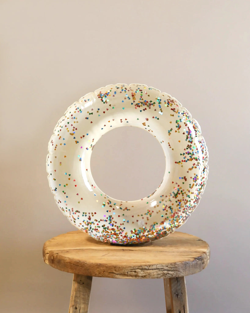 A large inflatable Junior Swim Ring with clear glitter hearts design sits on an old wooden stool against a plain light gray background.