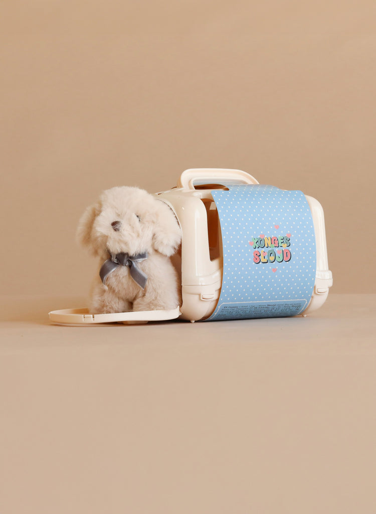 A cuddly Konges Sloejd Take Care of Me Animals plush dog sits inside a small white plastic pet carrier with a blue polka-dot door, waiting for someone to look after it.