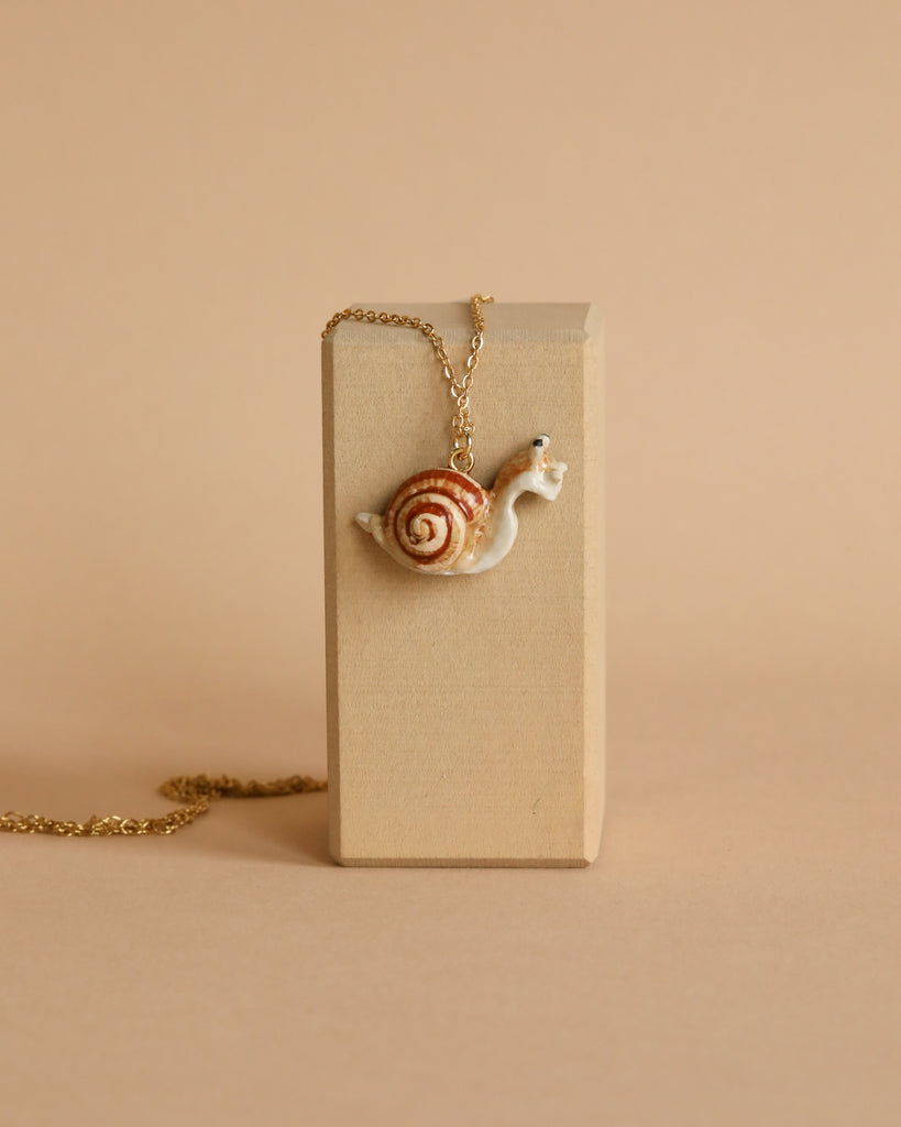 A delicate Snail Necklace with a snail-shaped pendant featuring a white body and brown spiral shell draped over a beige rectangular block against a matching beige background. The 24k gold-plated chain of the handmade nature necklace adds heirloom quality and elegance to the whimsical design.