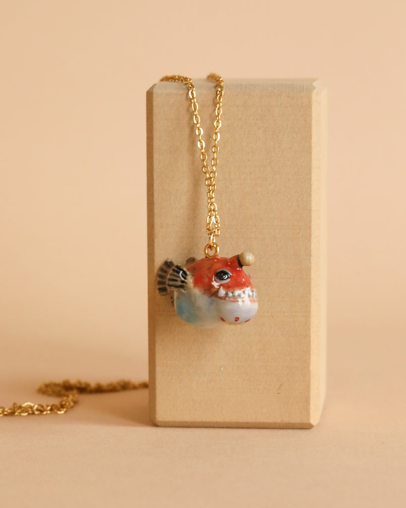 A gold-plated chain necklace with a pendant shaped like an angler fish rests on a rectangular wooden block set against a light beige background. The handcrafted porcelain pendant features a red and white body with black and white fins and detailed facial features.