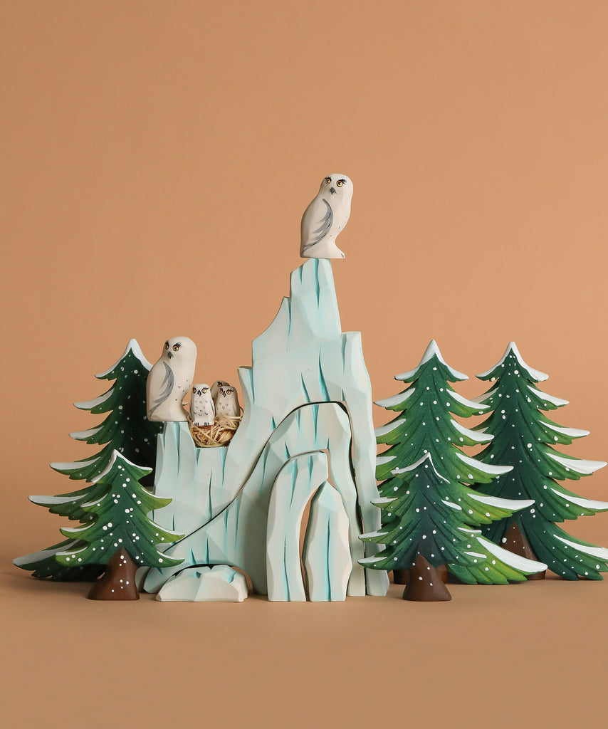 The Bumbu Handmade Wooden Snowy Owl Wilderness Set features snowy owl and owlet figurines on a light blue mountain with green pines, ideal as a Montessori educational toy displayed against a beige background.