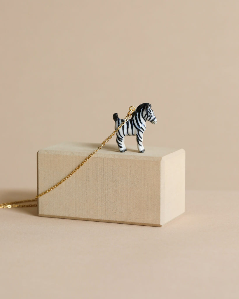 A Zebra Necklace is displayed on a light wooden block against a beige background. The zebra charm is black and white with delicate features, and the gold chain is partially visible extending beyond the block.