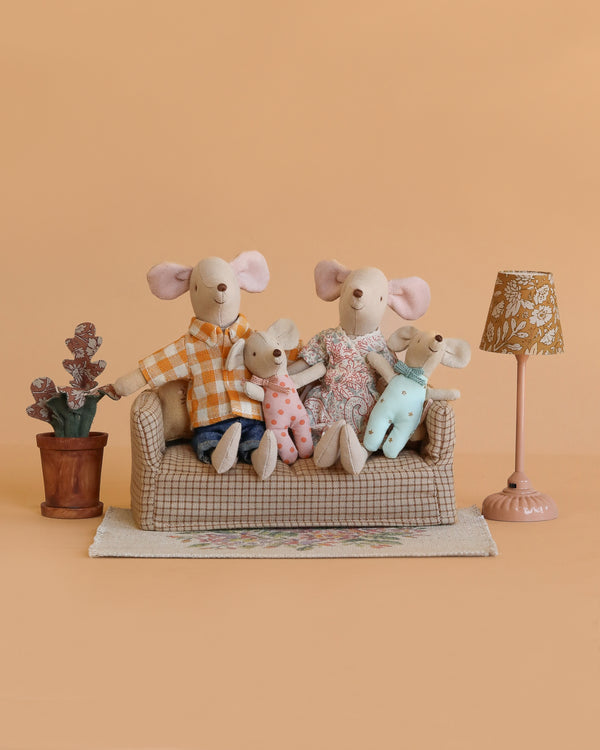 The Maileg Family Living Room Set features four dressed mice on a small couch, with a potted plant, a beige flower rug, and a patterned lamp, all set against a light brown background.