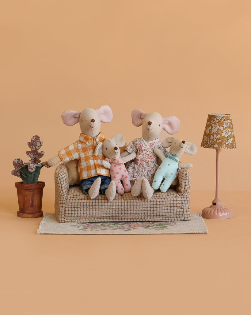 The Maileg Family Living Room Set features four dressed mice on a small couch, with a potted plant, a beige flower rug, and a patterned lamp, all set against a light brown background.