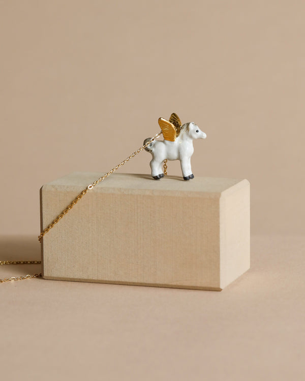 Sentence with given product name: A small, winged Gold Pegasus Necklace on a delicate 24k gold plated chain rests atop a beige jewelry box against a soft beige background.