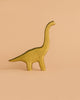 A yellow dinosaur from the Handmade Holzwald Dinosaur Set, crafted from wood and resembling a brontosaurus, stands on a simple peach background.