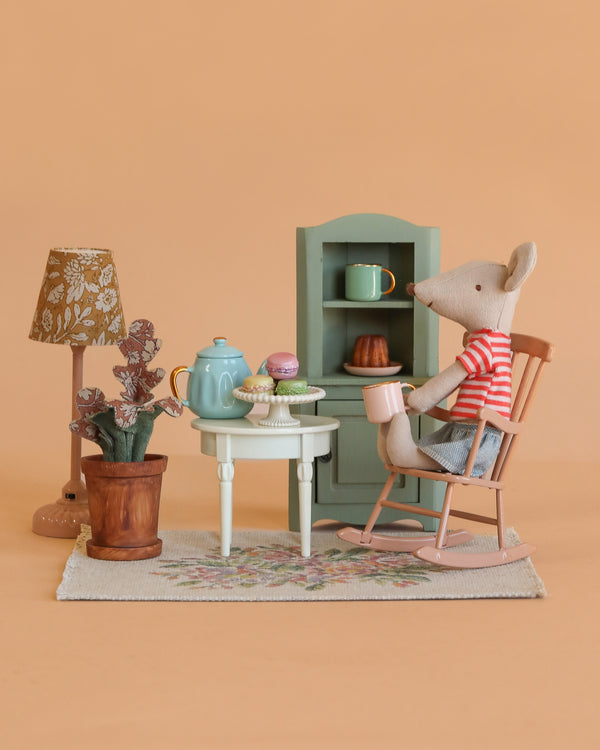 A Maileg Big Sister Afternoon Tea Set features a mouse in a striped shirt sitting in a rocking chair at a small table with tea, pastries, and flowers, surrounded by miniature furniture against a peach background.