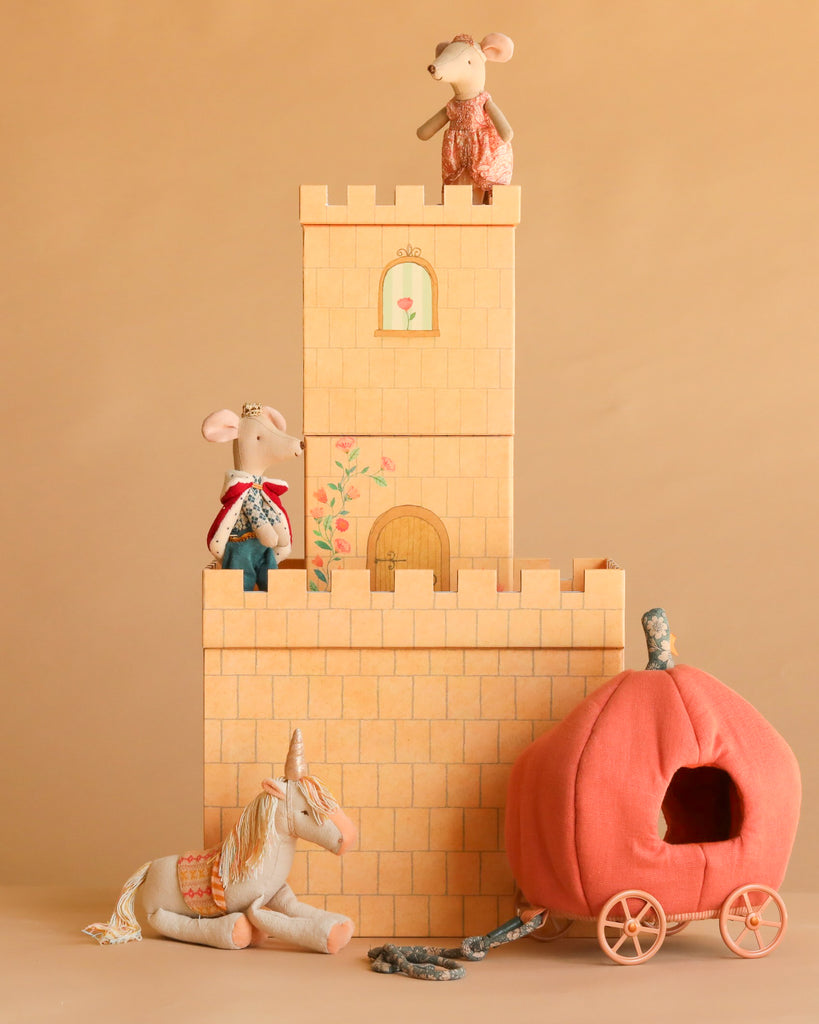 The Maileg Fairytale Set, featuring three toy mice on a Maileg Castle, a plush unicorn, and an orange pumpkin carriage, is displayed against a tan background to create a charming fairytale scene.