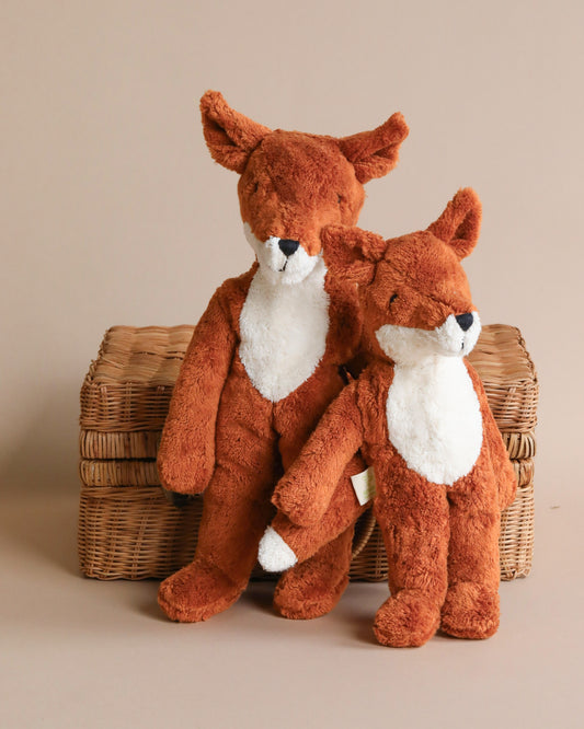 Two Senger Naturwelt Stuffed Animal - Foxes sitting snugly next to each other on a wicker basket, against a neutral beige background. The foxes are in a sitting position and appear soft and cuddly.