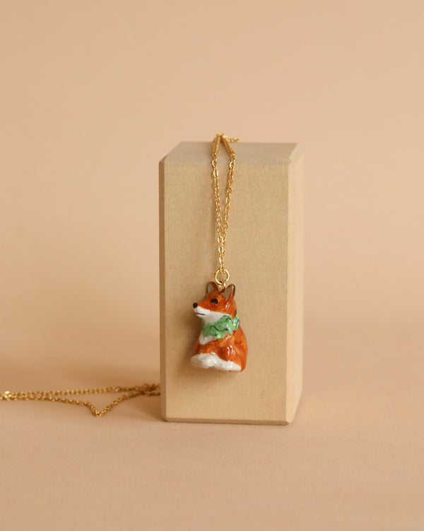 A Fox Necklace with a pendant shaped like a small corgi dog sitting upright, wearing a green scarf, is displayed draped over a rectangular beige block against a beige background. The hand-painted details on the gold-plated chain highlight its charming design.