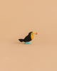 The Handmade Holzwald Toucan Bird is a small, colorful wooden figurine with a yellow chest, black body, blue feet, and a red-green beak—an eco-friendly piece for any sustainable toy collection, shown against a plain beige background.