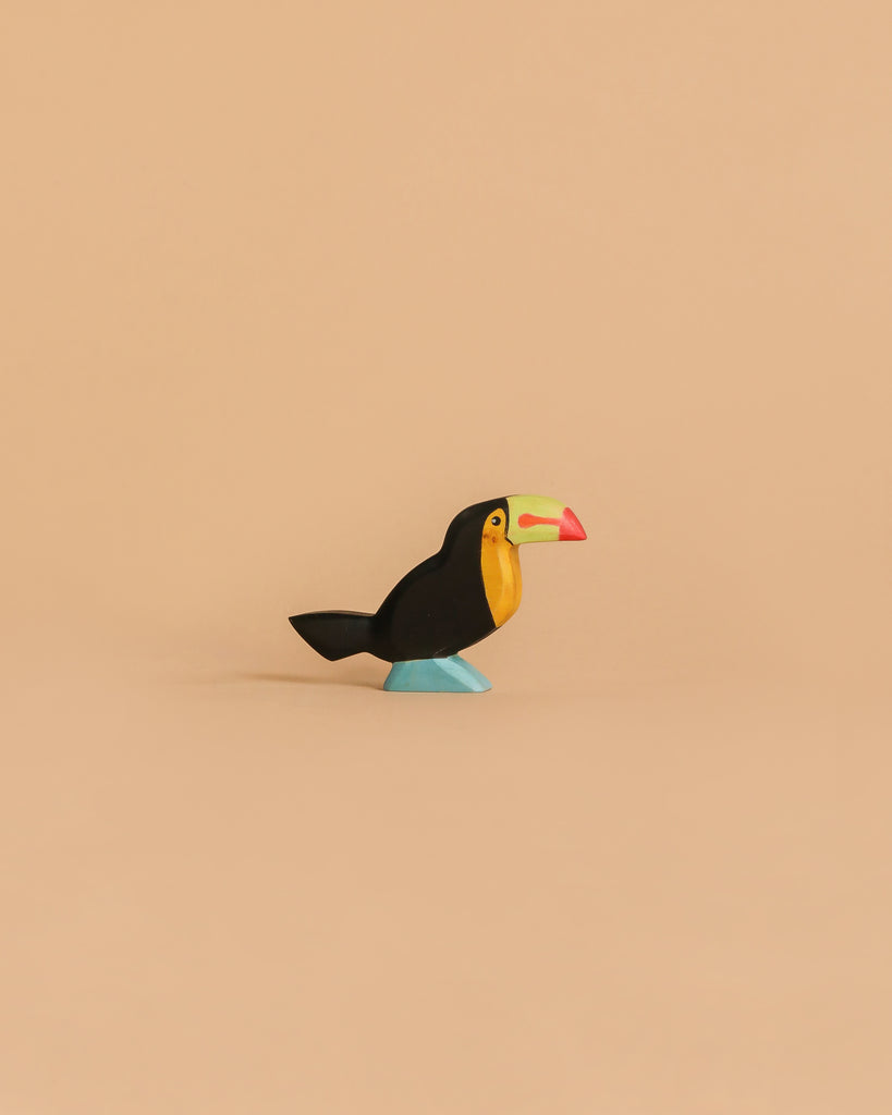 The Handmade Holzwald Toucan Bird is a small, colorful wooden figurine with a yellow chest, black body, blue feet, and a red-green beak—an eco-friendly piece for any sustainable toy collection, shown against a plain beige background.