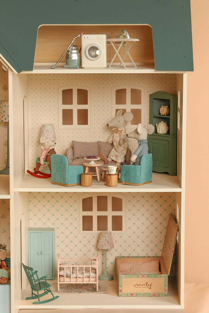 The Maileg Fully Furnished Dollhouse Set + Surprise Gift features three rooms: a laundry with washer and iron, a living room with sofas, two mice, lamp, and a nursery with crib, rocking chair, and toy chest.