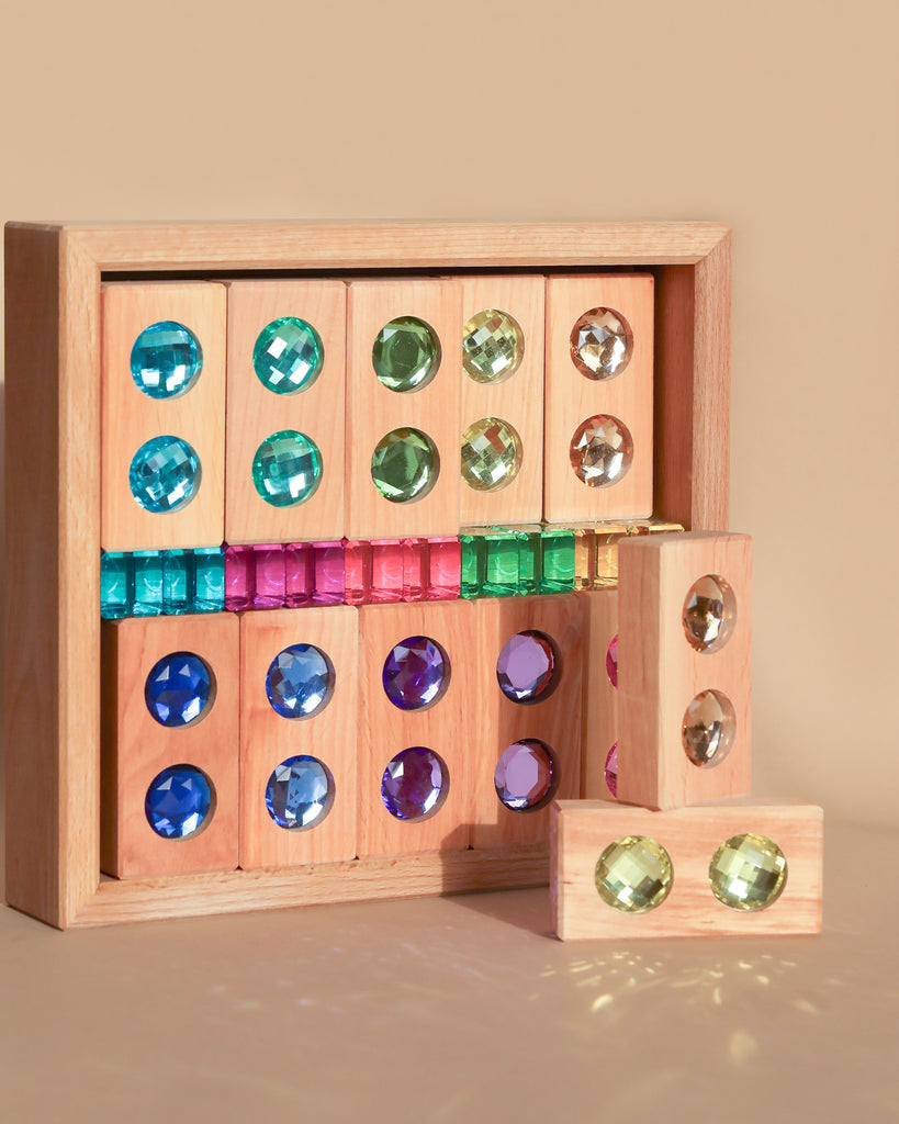 The Bauspiel Gem Blocks with Pastel Lucent Cubes (45 pieces) set includes wooden blocks adorned with colorful gems, creating a lively grid in shades of blue, green, purple, and pink on the blocks within and around a wooden frame against a beige background.