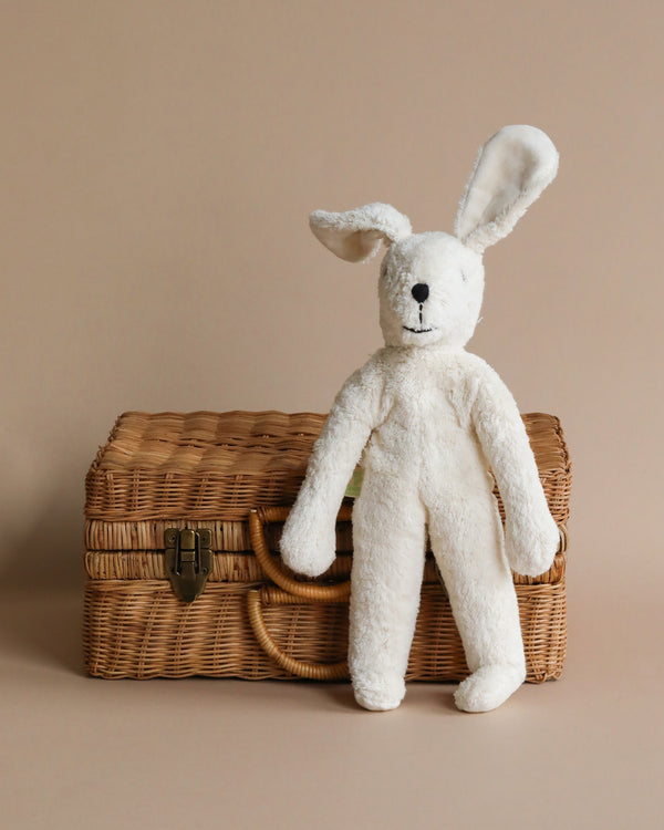 A Senger Naturwelt Stuffed Animal - White Bunny made from organically grown cotton leaning against a closed wicker basket on a plain beige background. The bunny has long floppy ears and a soft, textured surface.