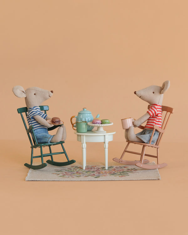 The Maileg Big Brother and Sister Afternoon Tea Set features two mice in striped shirts enjoying tea and pastries at a small table on a floral rug, with Maileg rocking chairs, set against a peach-colored background.